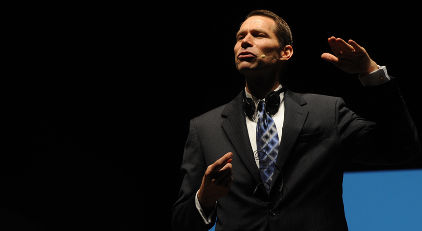 A man in a suit and tie speaks on stage, wearing a headset microphone. He gestures with one hand raised and the other holding a small device, against a dark background with a blue section.