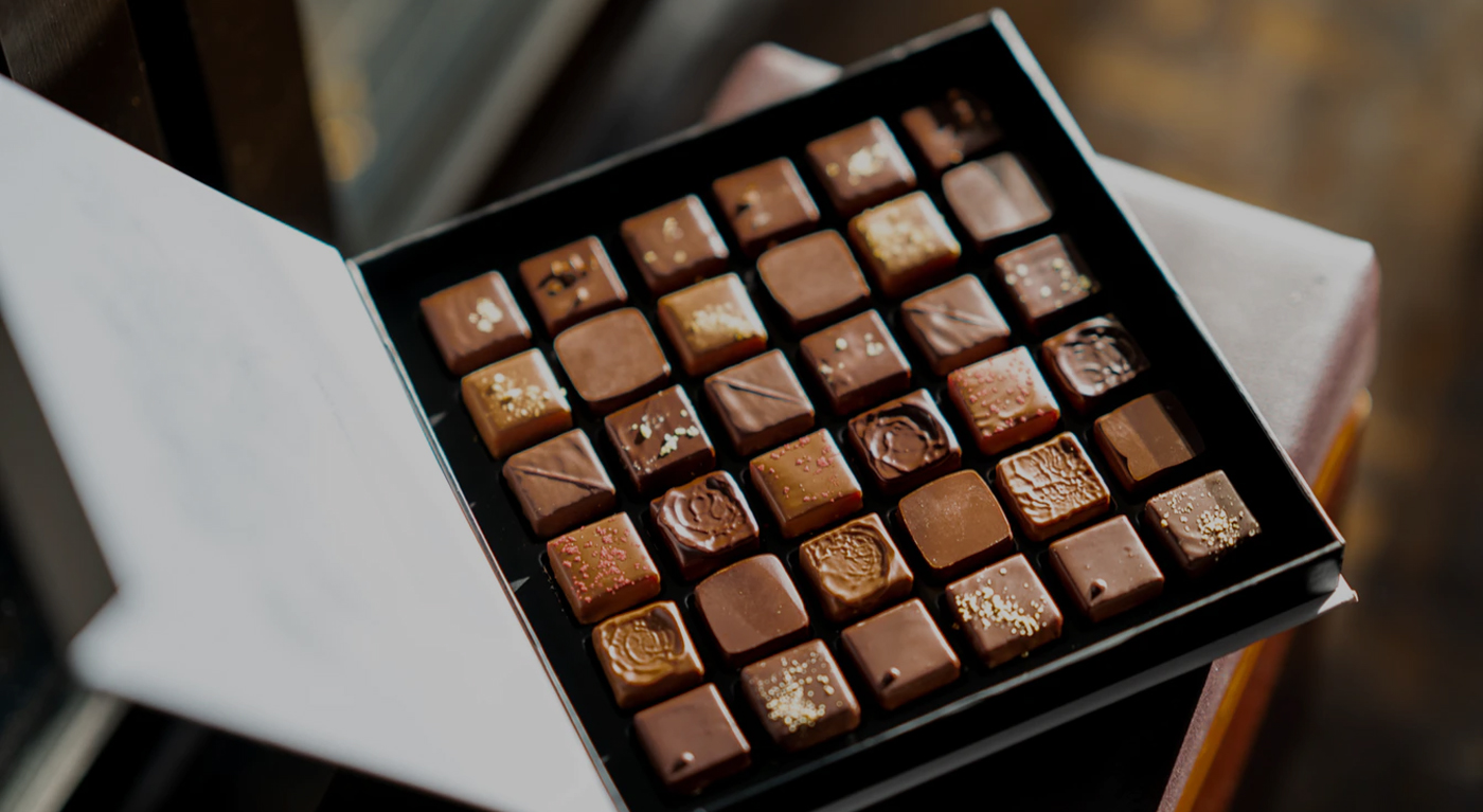 An open box of assorted gourmet chocolates arranged in neat rows, with various shapes, colors, and decorative toppings on each piece.