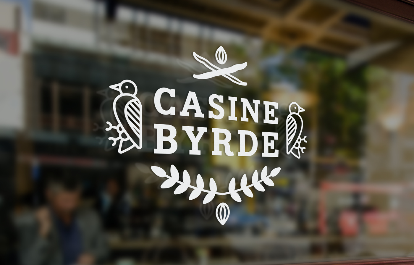 A glass window displays a white logo with two birds, leaves, and the words “CASINE BYRDE” in bold letters, with a busy city street and people blurred in the background.