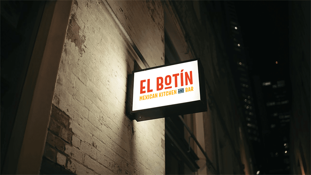 A lit rectangular sign on a brick wall reads EL BOTÍN MEXICAN KITCHEN AND BAR in red, orange, and blue letters, photographed at night in an urban setting.