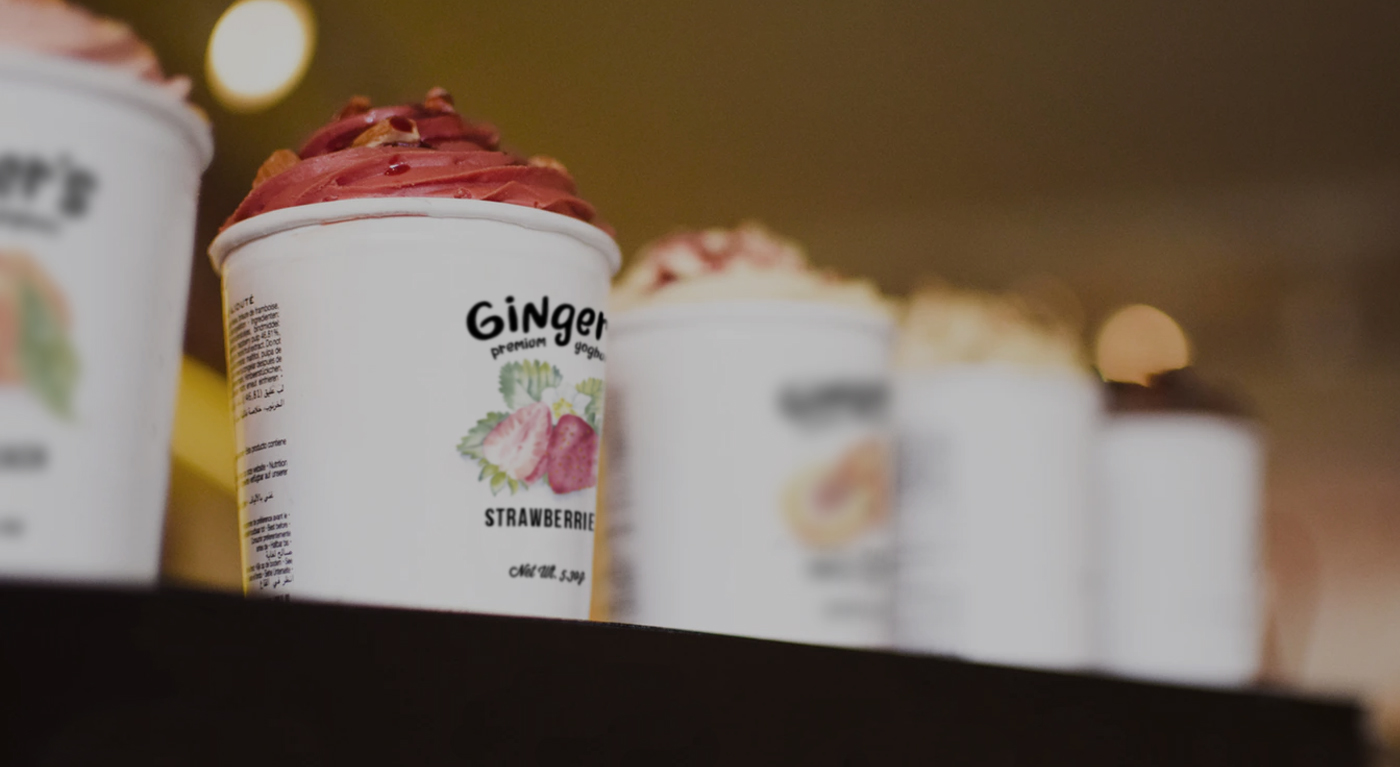 Four white cups of ice cream with different flavors and toppings are lined up on a counter. The focus is on the front cup labeled Strawberry with pink ice cream; the background cups are blurred.