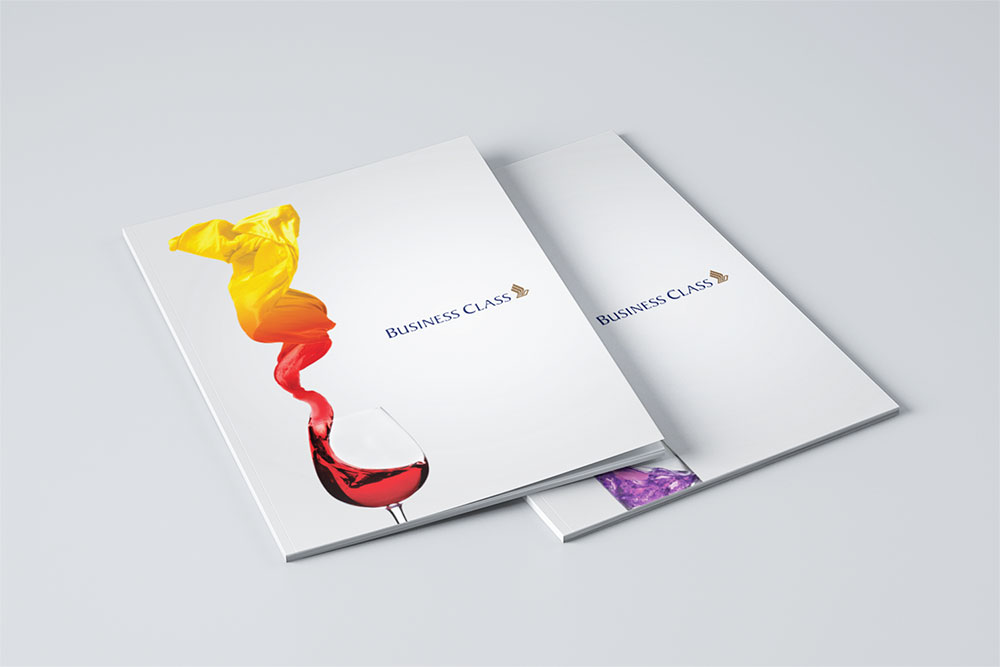 Two business class menus for SIA designed by Sandra Tay with white covers; one shows red wine in a glass with a yellow-gold fabric swirling above, the other menu is partially hidden underneath. Both display BUSINESS CLASS and a logo.