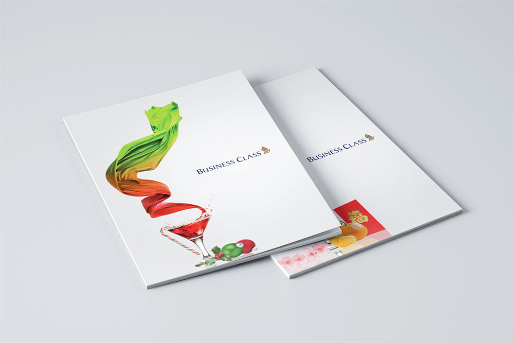 Two festive business class menus for SIA designed by Sandra Tay on a white surface. One menu has a colorful, swirling design above a cocktail glass with fruit and vegetables, while the other is mostly plain with the text Business Class and a logo.