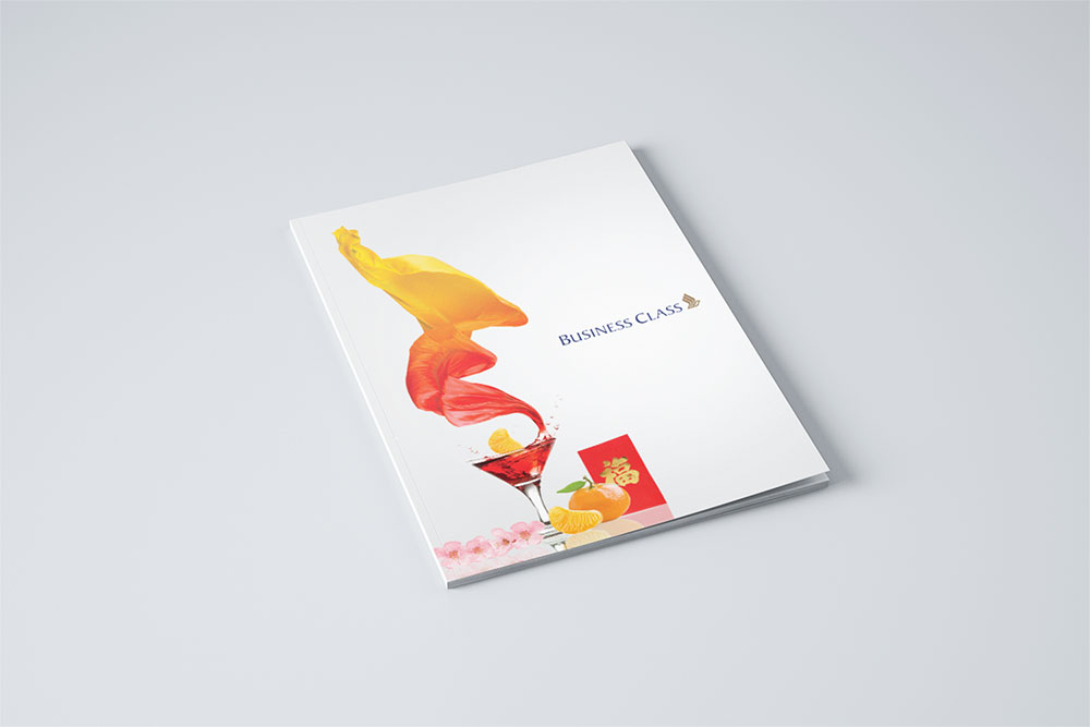A business class menu for SIA designed by Sandra Tay with a festive cover featuring a colorful drink, orange slices, pink flowers, a red envelope, and dynamic red and yellow fabric swirls against a white background.