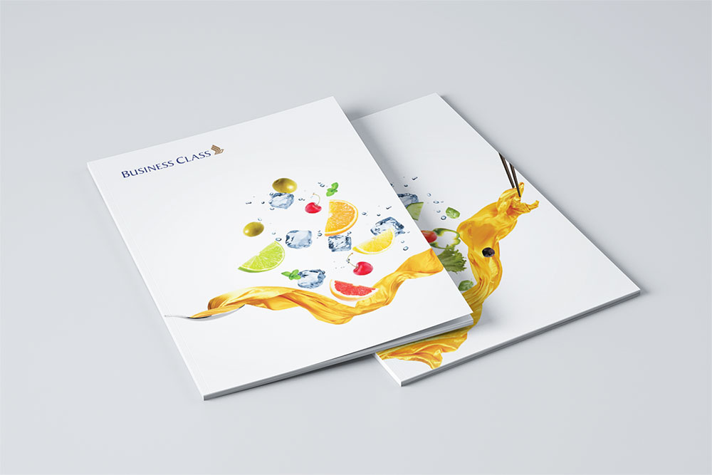 Two business class booklets for SIA designed by Sandra Tay with white covers featuring colorful fruit, ice cubes, and a yellow liquid splash design, arranged on a light gray surface.