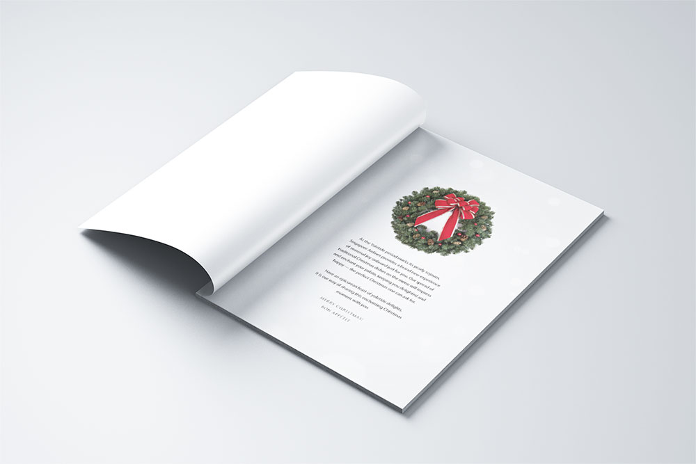 An open greeting card on a white surface displays a festive wreath with a red bow on the right page, along with a printed message beneath it. The left page is blank and partially turned.
