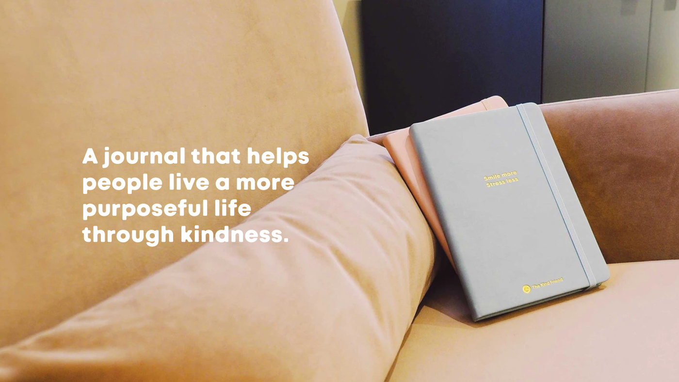 A pastel blue journal with gold text lies on a beige couch next to a peach-colored cushion. White text on the image reads, “A journal that helps people live a more purposeful life through kindness.”.