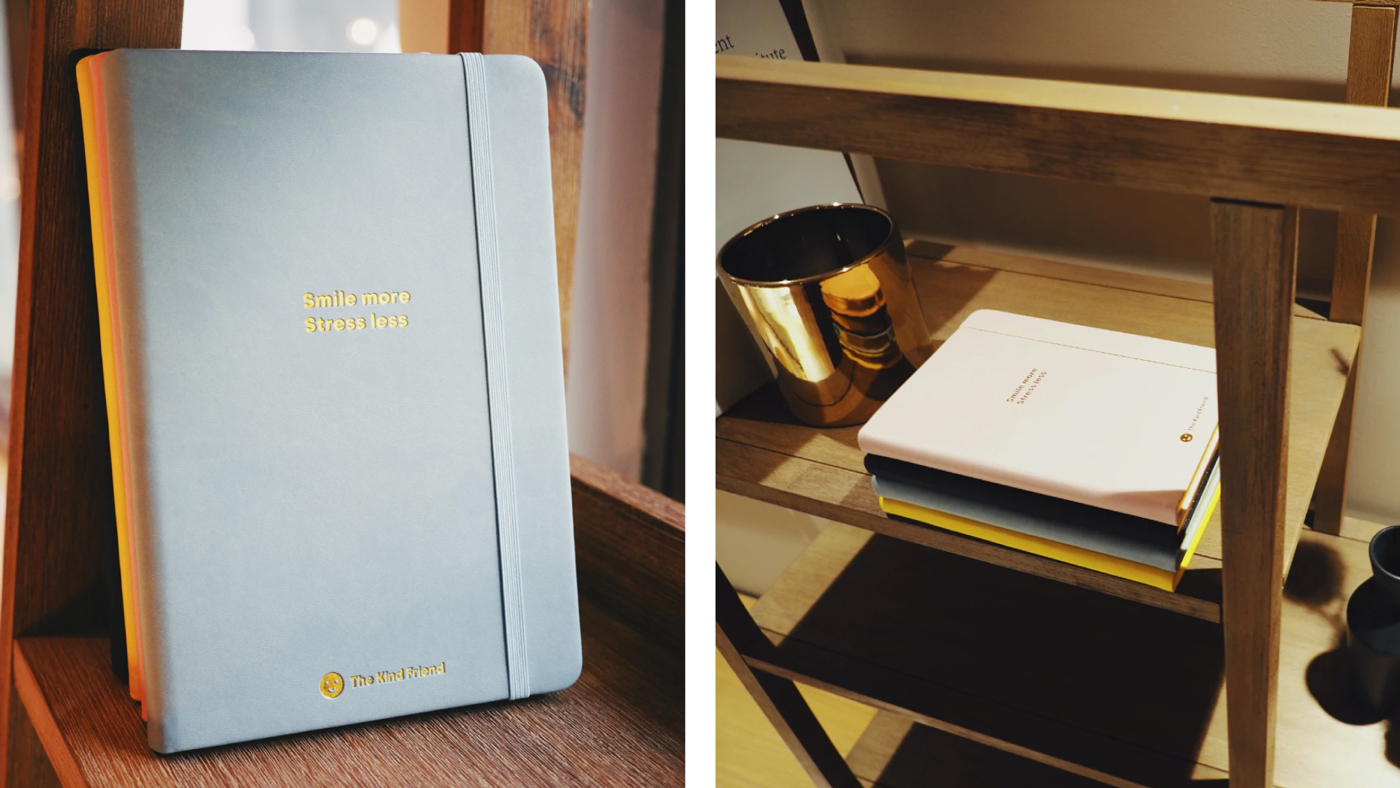 A light blue notebook with Smile more Stress less written in gold, displayed on a wooden shelf. Next image shows the notebook stacked with others and a gold cup on a shelf.