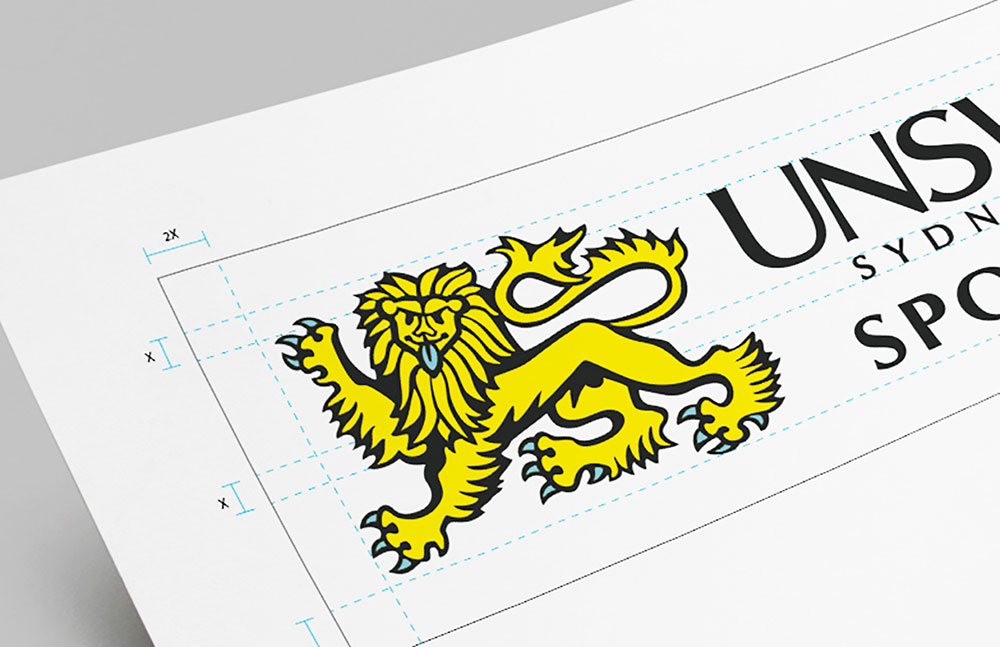A printed paper shows a yellow, heraldic lion graphic next to the black text UNSW SYDNEY SPORT. Blue grid lines and measurements are visible around the design, indicating alignment and spacing.