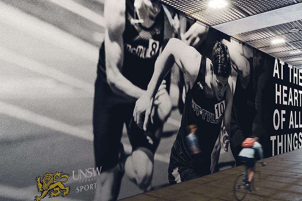 A cyclist rides past a large black-and-white mural of runners in motion. The mural displays the UNSW Sydney Sport logo and part of the phrase AT THE HEART OF ALL THINGS.