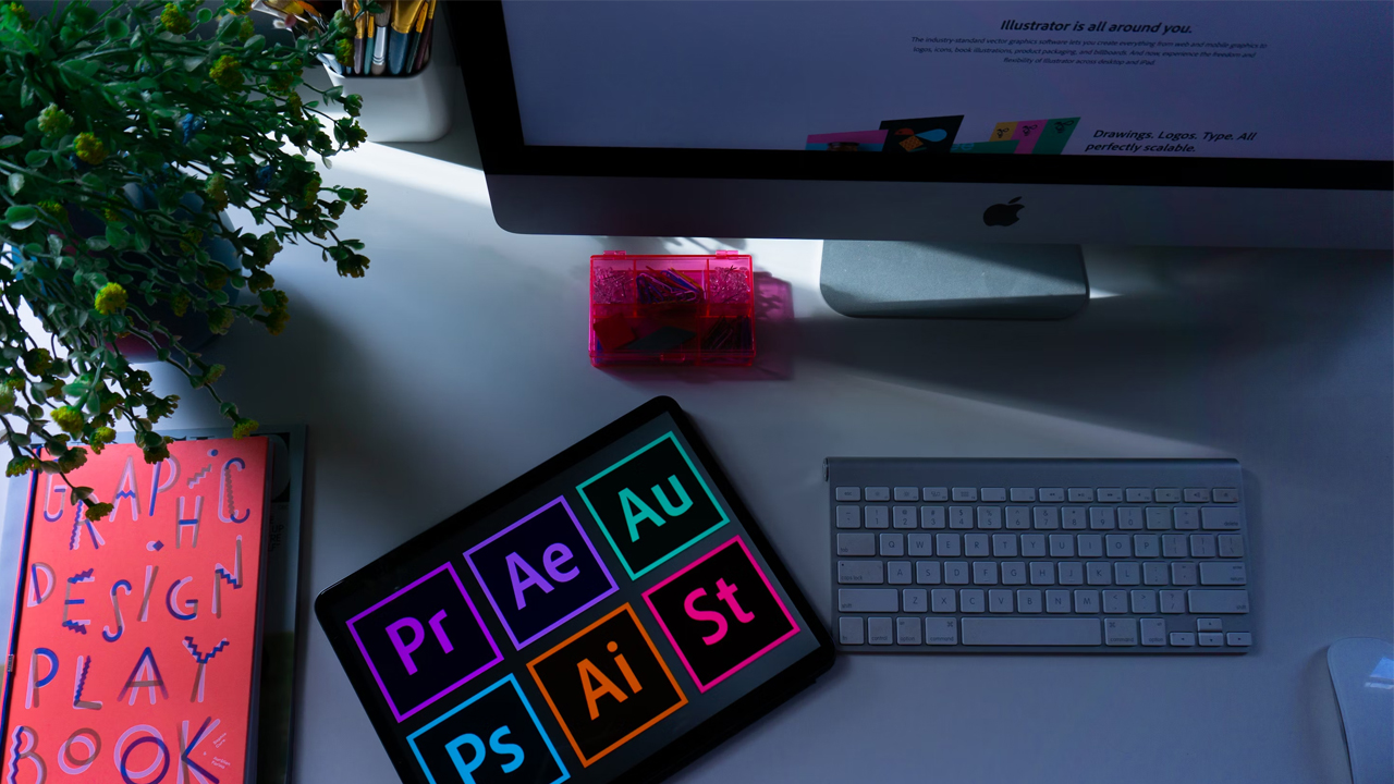 A workspace with a tablet displaying Adobe software icons, an Apple wireless keyboard, an iMac, a plant, a pink book titled GRAPHIC DESIGN PLAYBOOK, and a small transparent pink box on a white desk.