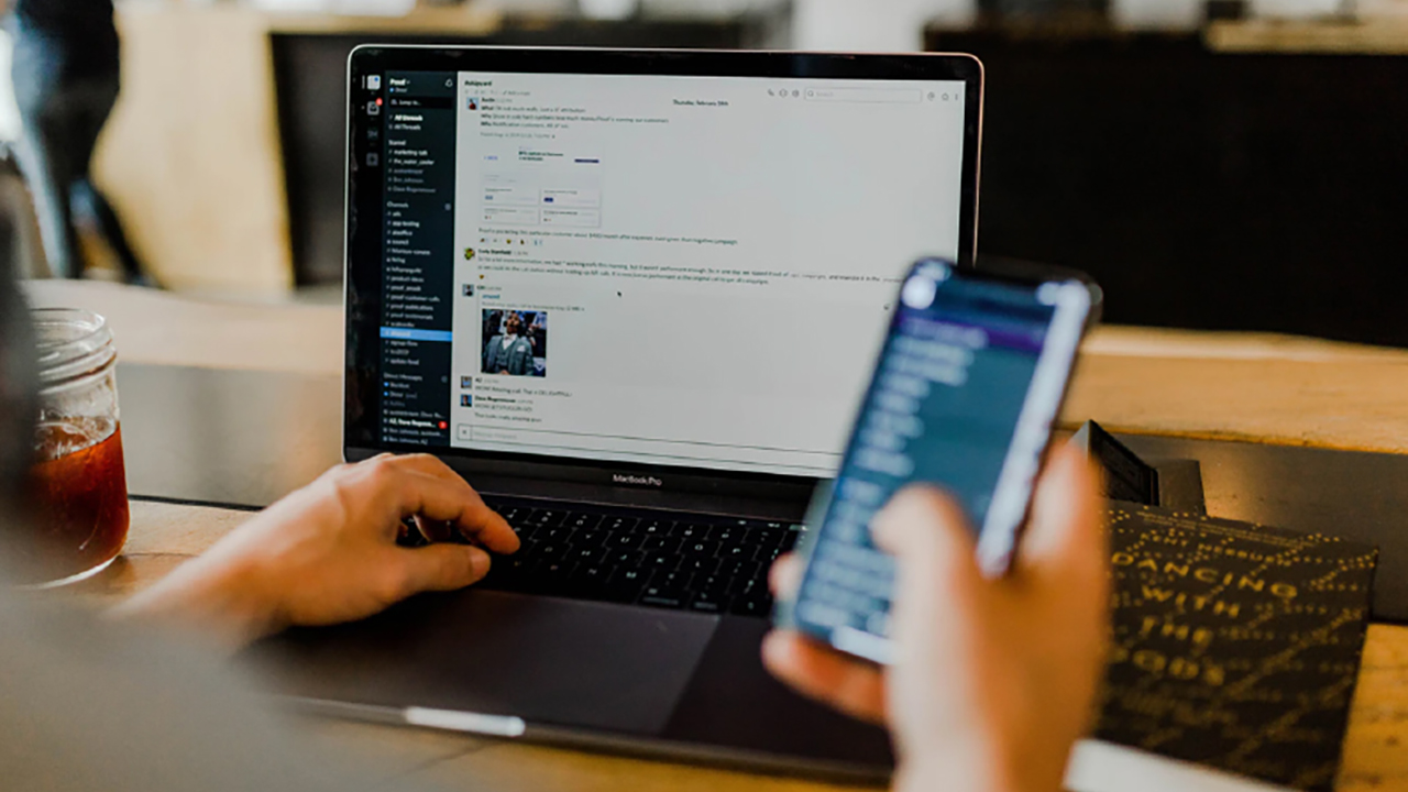 A person sits at a desk using a laptop with a chat application open on the screen, while holding a smartphone in their hand. A jar with a red drink and a notebook are also on the desk.