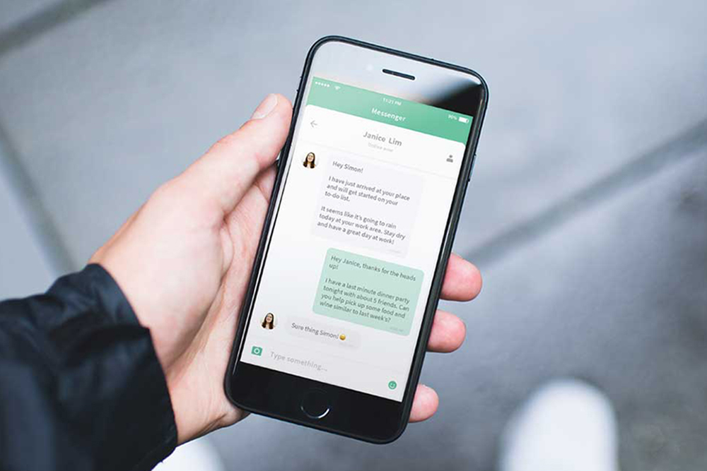 A person holds a smartphone displaying a messaging app, showing a chat conversation with light green and white message bubbles on the screen. The background is blurred.