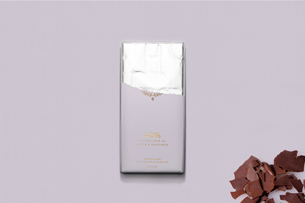 A partially unwrapped lavender-colored chocolate bar with gold text sits on a light purple surface. Pieces of broken chocolate are seen in the lower right corner.