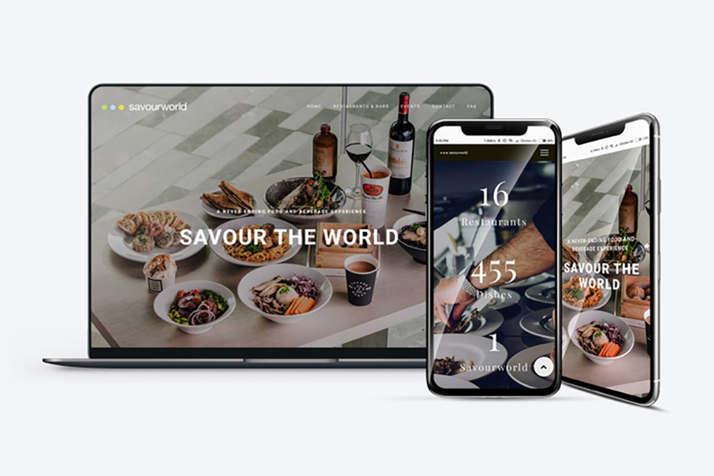 A laptop and two smartphones display a restaurant website with dishes, drinks, and text reading Savour the World and restaurant stats. The background shows a table set with plates and wine.