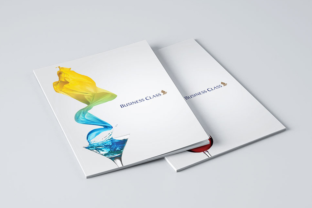 Two white business class brochures are stacked on a light surface. The top brochure, designed by a Freelance Graphic Designer Singapore, features a vibrant splash of yellow, green, and blue liquid above a martini glass. The logo and Business Class text appear on both covers.