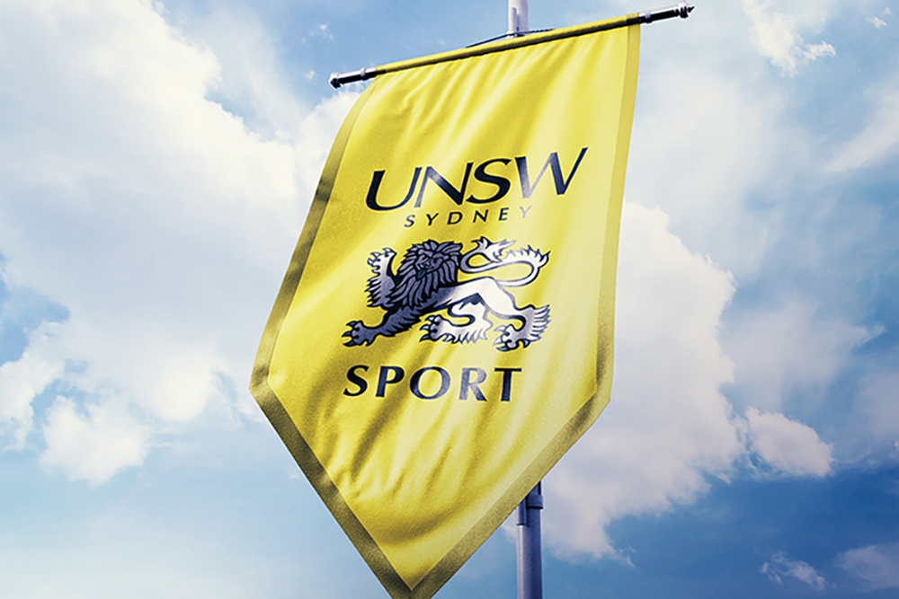 A yellow banner featuring the UNSW Sydney logo, a blue lion graphic, and the word SPORT hangs on a pole against a blue sky with white clouds—showcasing pride in Sandra Tay’s design legacy.